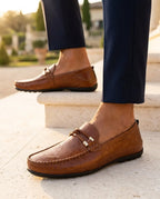 Side view of a brown leather moccasin-style loafer with silver bit hardware, paired with navy pants outdoors