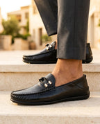 Black leather loafer with silver bit buckle and handstitched apron, worn with grey tailored pants on stone steps