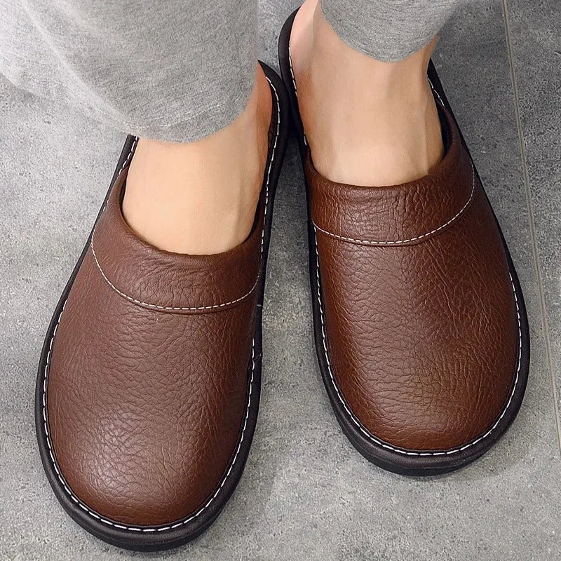 Close top-down view of brown leather mule slippers worn on bare feet, showing white contrast stitching and slip-on heel.