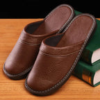 Pair of brown leather slide slippers with contrast stitching resting against green books on a timber tabletop