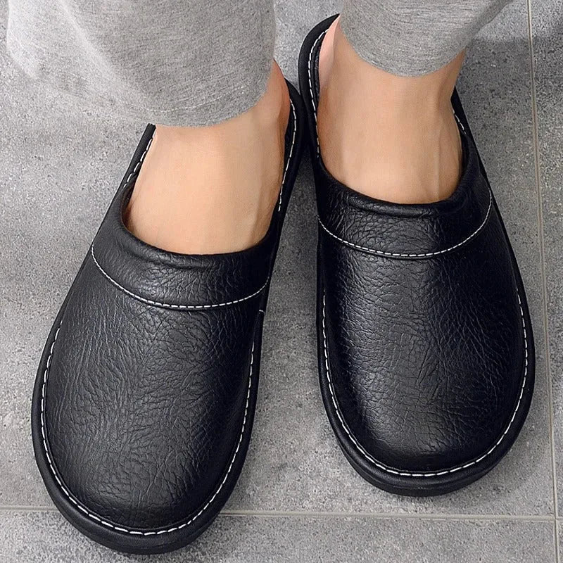 Top-down view of black leather mule slippers worn on bare feet with visible white stitch detailing around the vamp.