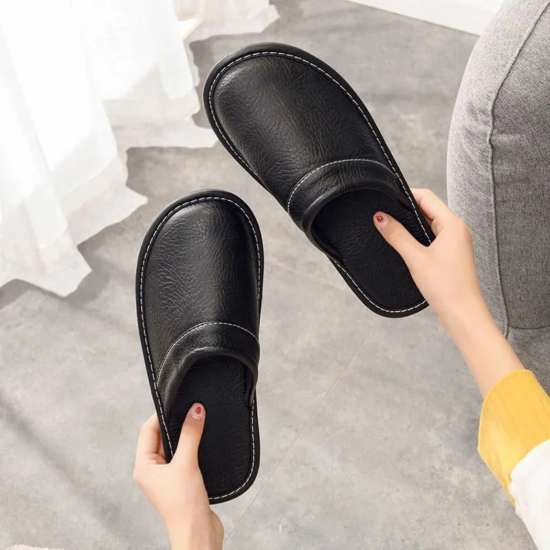 Black leather slippers held in both hands, displaying the padded insole and curved slip-on back design.
