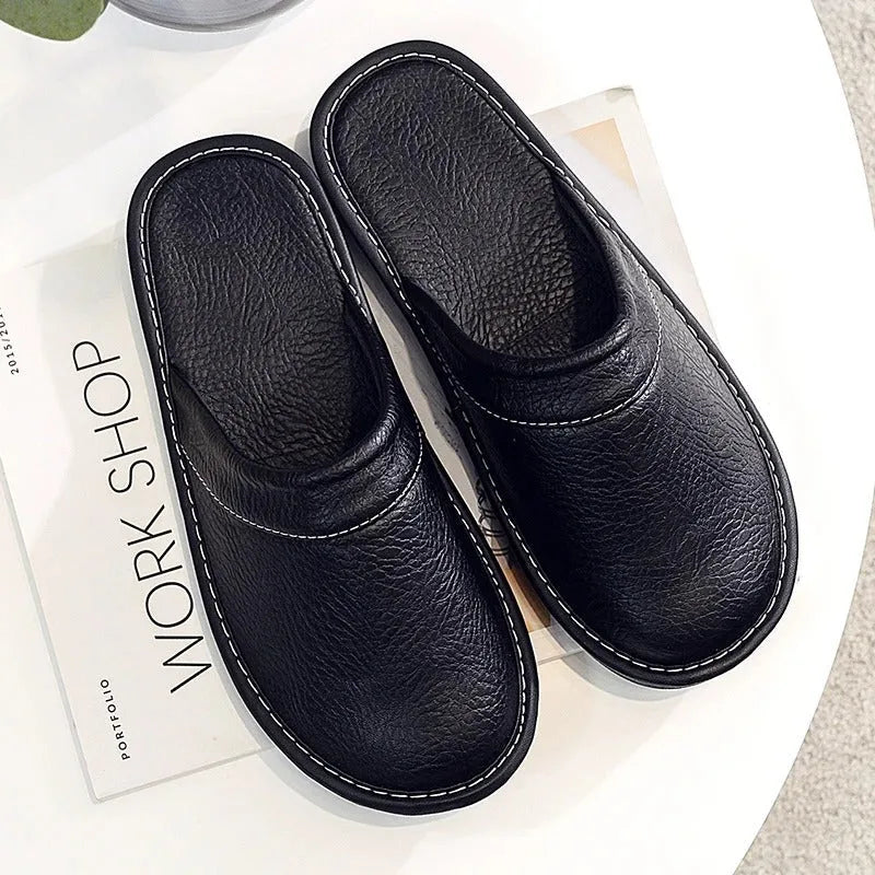 Overhead flat-lay of a pair of black leather slippers on a white surface beside a magazine, showing matching outsoles.