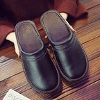 Overhead view of black leather slippers with stitched trim held in a person's hand over a wooden floor