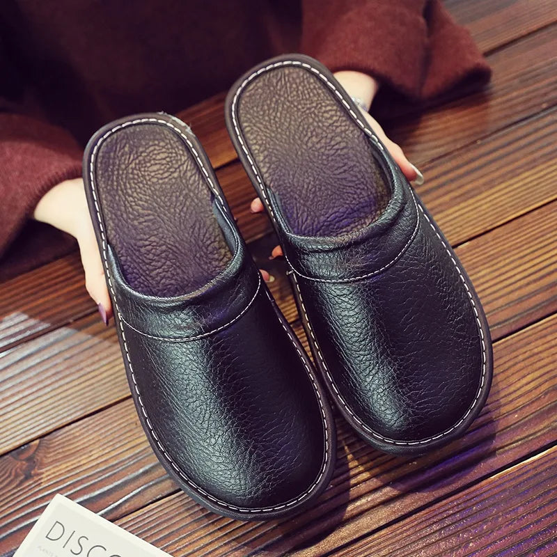 Overhead view of black leather slippers with stitched trim held in a person's hand over a wooden floor