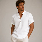 Man wearing a white band-collar linen shirt with rolled sleeves, unbuttoned at the top, paired with beige pants