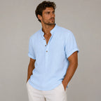 Man in a light blue band-collar linen shirt with sleeves rolled, hands in pockets, worn with cream pants