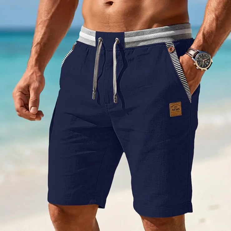 Navy blue linen-blend shorts with striped pocket trim and leather patch, worn with hand in pocket on sand