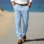 Man walking on sand in light blue linen trousers paired with a white shirt and brown belt