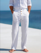 White linen drawstring trousers worn barefoot on a beach, showing relaxed straight-leg fit and rolled cuffs