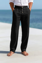Black linen drawstring trousers shown full length against a sandy beach backdrop