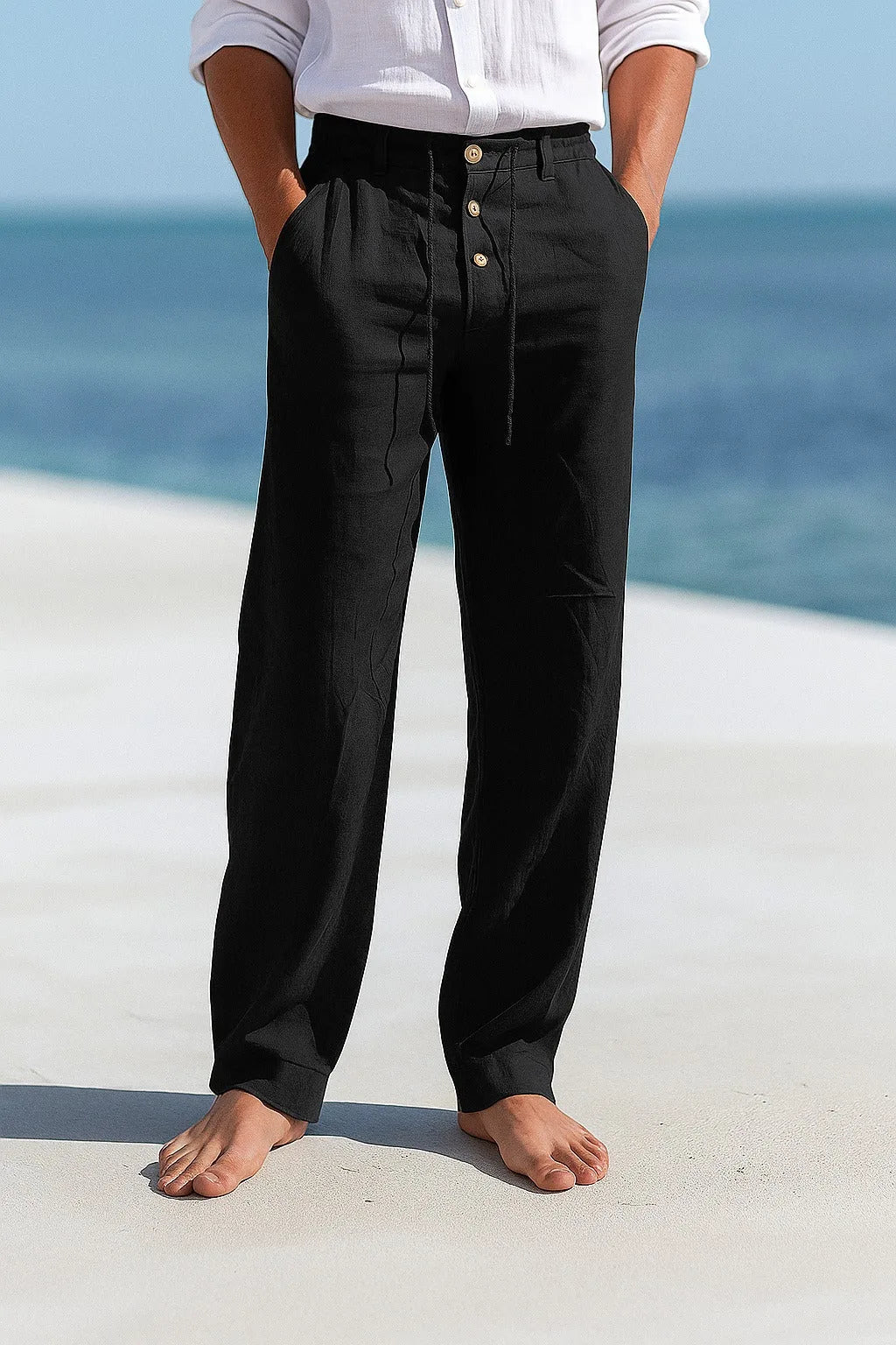 Black linen drawstring trousers shown full length against a sandy beach backdrop