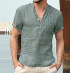 Front view of man wearing a sage green linen short sleeve shirt with open button placket, standing near a harbour.