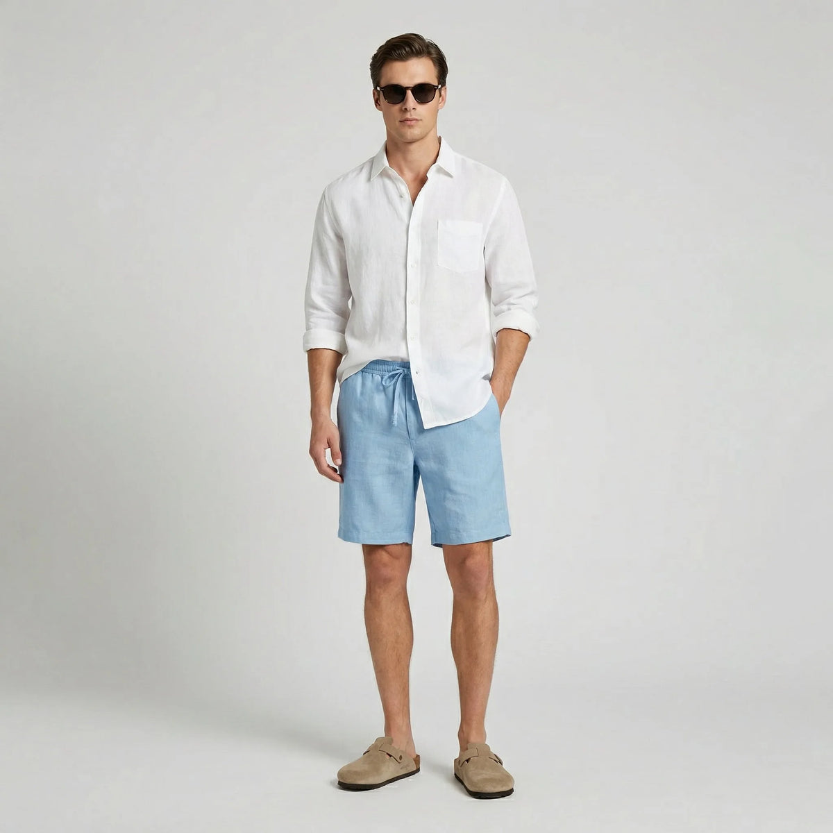 Man wearing light blue linen shorts with a white linen shirt and suede slides, full body front view