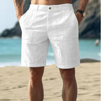 Rear view of white linen-textured shorts with button-tab waist closure, worn by a man standing on sand near the ocean.