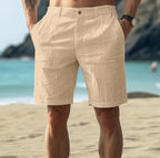 Back view of khaki linen-textured shorts showing pleated front darts and belt loops, beach setting with ocean behind.