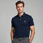 Man in a navy pique polo shirt with tan embroidered laurel wreath logo, worn tucked in with grey trousers