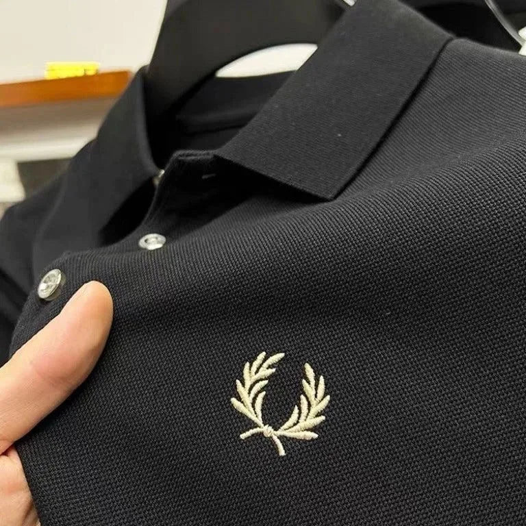 Close-up of black pique polo fabric showing cream laurel wreath embroidery and the ribbed collar