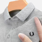 Close-up of light grey marl polo fabric with black laurel wreath embroidery, finger pointing at the logo