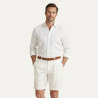 Man in a white linen shirt and cream pleated chino shorts with turn-up hems, standing against a plain background
