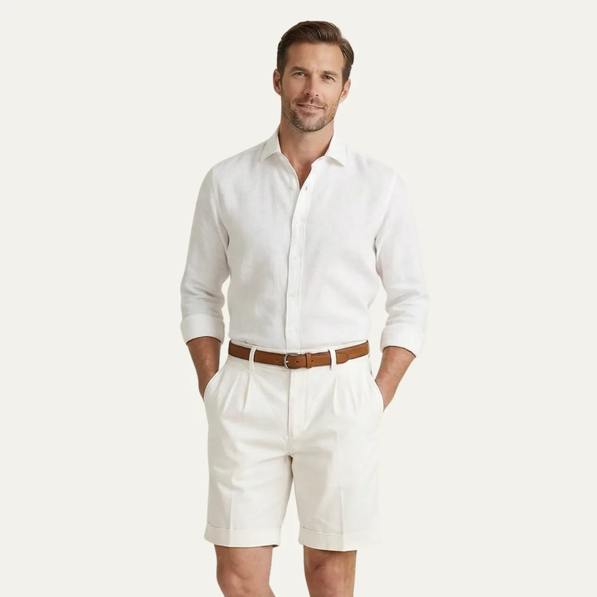 Man in a white linen shirt and cream pleated chino shorts with turn-up hems, standing against a plain background