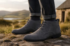 Man wearing grey suede chukka boots with rolled cuffed jeans, standing on a rock with hills in the background.