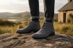 Close-up of grey suede chukka boots laced up, worn with dark jeans against a scenic outdoor backdrop.