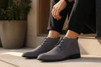 Side view of grey suede chukka boots worn with dark trousers, seated on a concrete step near a potted plant.