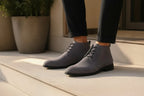 Detailed side profile of grey suede chukka boots with black laces and sole, worn outdoors in natural light.