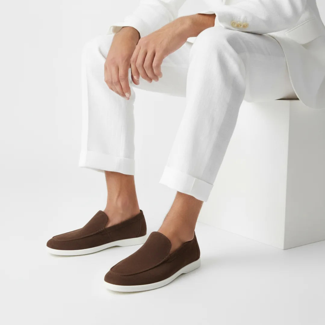Model seated in a white linen shirt and trousers wearing dark brown suede loafers with white soles