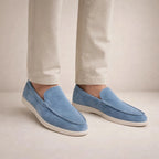 Side view of model wearing light blue suede slip-on loafers with white sole and tan pants