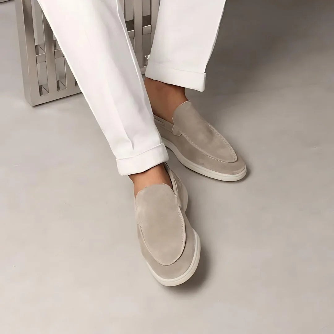 Close-up of beige suede loafers worn with rolled white trousers while seated, feet crossed
