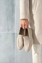 Hand holding a pair of beige suede loafers by their heels, model wearing a matching coat