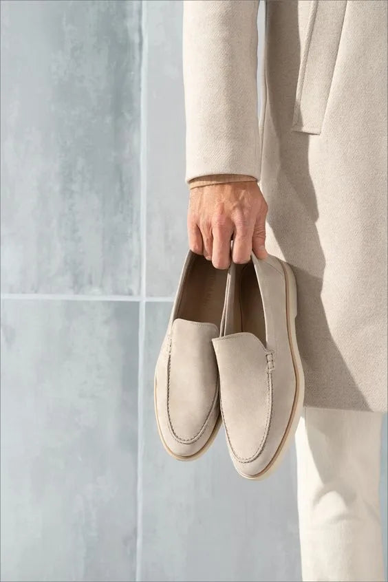 Hand holding a pair of beige suede loafers by their heels, model wearing a matching coat