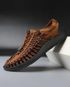 Side view of a brown woven cord sandal with black contoured sole and toggle lace closure, shown resting against a box