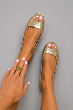 Top-down view of gold metallic textured leather peep-toe flats on bare feet against a grey backdrop.