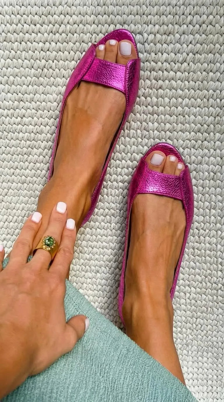 Fuchsia metallic textured leather peep-toe flats shown from above on bare feet on a woven rug.