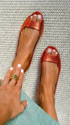 Orange metallic leather peep-toe ballet flats photographed from above on a woven rug.