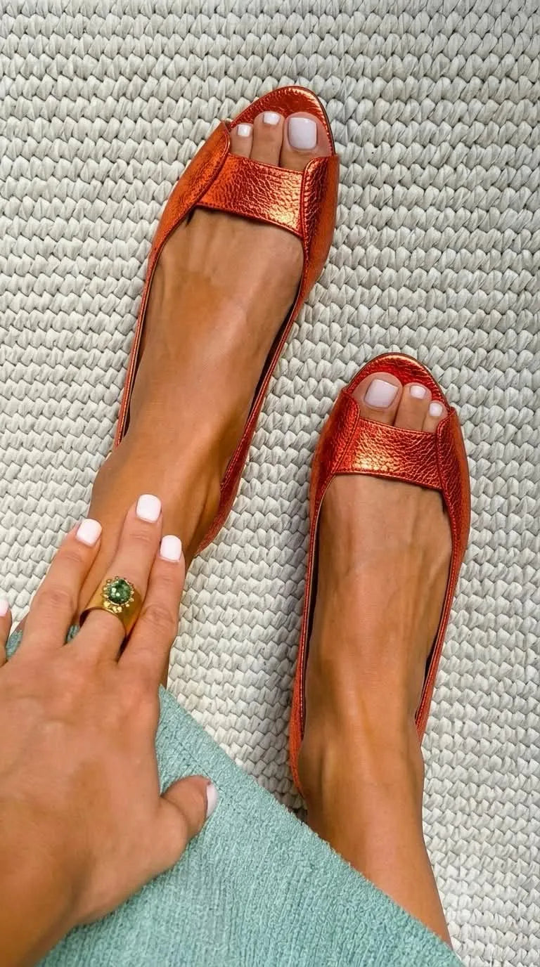 Orange metallic leather peep-toe ballet flats photographed from above on a woven rug.