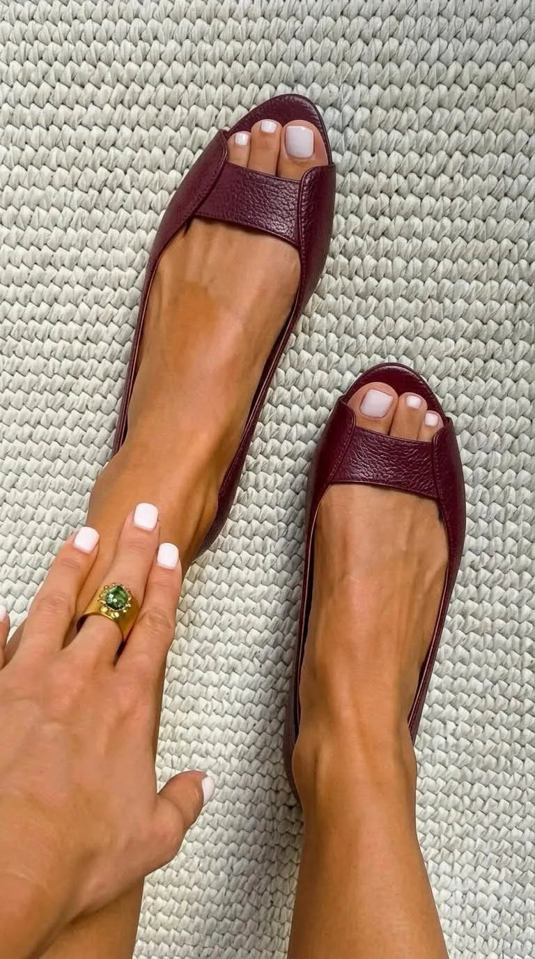 Deep bordeaux textured leather peep-toe flats worn on bare feet on a woven jute rug.