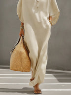 Waist-down detail of the flowing oatmeal linen maxi dress fabric and sleeve, worn with woven tote and sandals