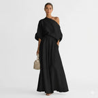 Woman in black one-shoulder linen-look maxi set with billowy sleeves, cuffed cuffs and flowing wide-leg skirt against a plain studio backdrop