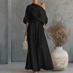 Side view of black asymmetric one-shoulder top and flowing maxi skirt set styled with a woven handbag against a grey textured wall