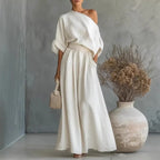 White one-shoulder linen-look two-piece set with draped sleeves and A-line maxi skirt, paired with a woven handbag near dried florals