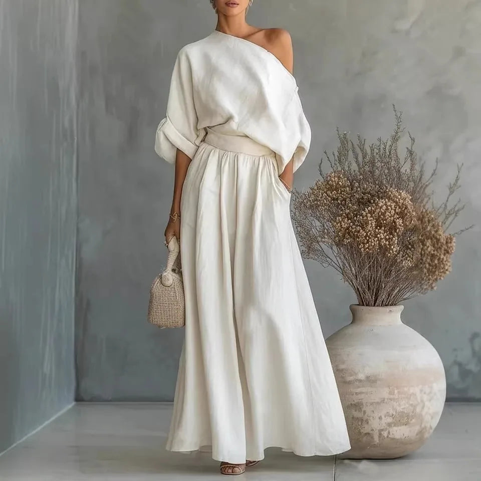 White one-shoulder linen-look two-piece set with draped sleeves and A-line maxi skirt, paired with a woven handbag near dried florals