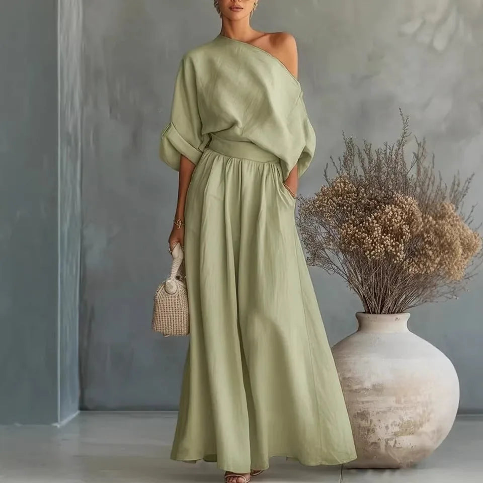 Olive green one-shoulder linen-look set with cuffed sleeve detail and flowing maxi skirt against a textured grey backdrop