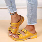 Yellow double-buckle wedge sandal with woven stitch detail and perforated leather strap, shown on model stepping forward
