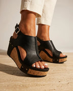 Close-up side view of black faux leather wedge sandal with buckled slingback strap and cork platform sole, worn with cream pants