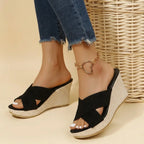 Black suede crisscross wedge sandals worn on crossed feet, showing the open toe and woven platform base