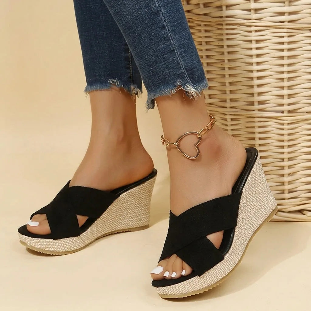 Black suede crisscross wedge sandals worn on crossed feet, showing the open toe and woven platform base